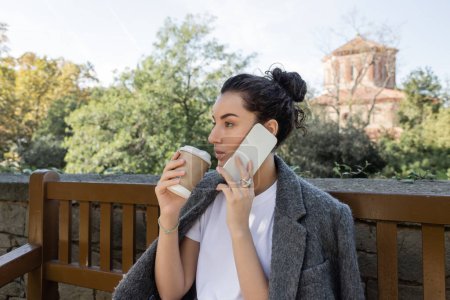 Hübsche und lockige Frau in T-Shirt und warmer Jacke, die am Smartphone spricht und Kaffee to go hält, während sie tagsüber auf einer Holzbank im Park in Barcelona sitzt, Spanien, Pappbecher mit Heißgetränk 