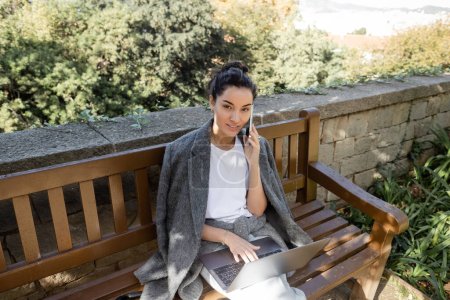 Mujer joven sonriente con una chaqueta cálida hablando en un teléfono inteligente y mirando a la cámara mientras trabaja en una computadora portátil y se sienta en un banco de madera en el parque en Barcelona, España, trabaja desde cualquier lugar 