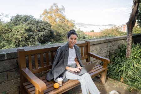 Joven freelancer sonriente con chaqueta abrigada y ropa casual mirando a la cámara, usando un portátil y sentado cerca del café para ir y naranja en un banco de madera en el parque en Barcelona, España 