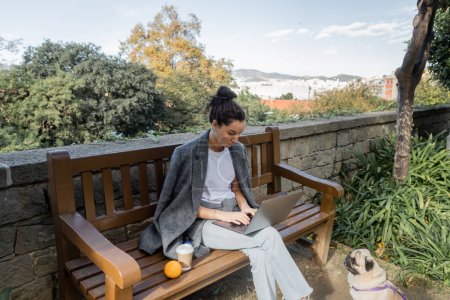 Joven freelancer morena en chaqueta abrigada trabajando en portátil cerca de café para ir y naranja fresca en banco de madera y perro pug sentado en el parque en Barcelona, España, durante el día 
