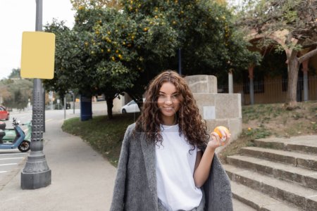 Mujer joven y rizada sonriente con chaqueta cálida sosteniendo naranja fresca y mirando a la cámara mientras está de pie en la calle urbana borrosa al fondo en Barcelona, España, farola, scooter