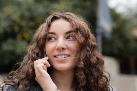 Photo for Portrait of young curly and brunette woman smiling and looking away while talking on smartphone and standing on blurred urban street in Barcelona, Spain, at daytime - Royalty Free Image