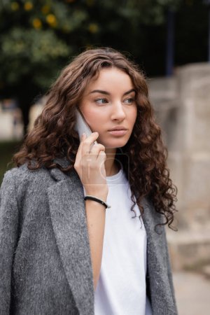 Portrait of pretty brunette and stylish young woman in warm jacket talking on cellphone and looking away while standing on blurred urban street in Barcelona, Spain 