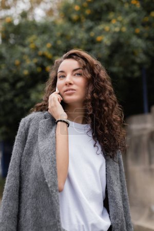 Joven mujer bonita y rizada con elegante chaqueta casual hablando en smartphone y mirando hacia otro lado mientras está parada en la borrosa calle urbana de Barcelona, España 