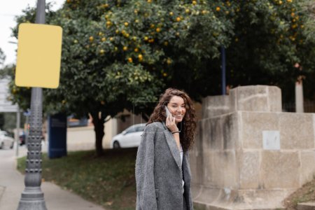 Carefree young curly woman in stylish casual jacket looking at camera while talking on smartphone and standing on urban street with street lamp and trees in Barcelona, Spain, at daytime 