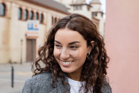 Portrait of smiling and curly woman in stylish casual jacket looking away while standing near blurred buildings at background on urban street in Barcelona, Spain 