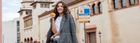 Young curly woman in casual grey jacket holding ripe and fresh orange and smiling at camera with historical landmark at background outdoors in Barcelona, Spain, banner, ancient building 