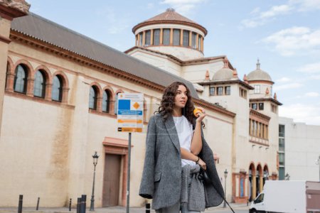 Young curly woman in grey casual jacket holding fresh orange and leash while looking away with blurred historical landmark at background in Barcelona, Spain, ancient building 