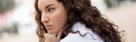Portrait of young and curly thoughtful woman in casual clothes holding hand near chin and looking away on blurred urban street in Barcelona, Spain, banner
