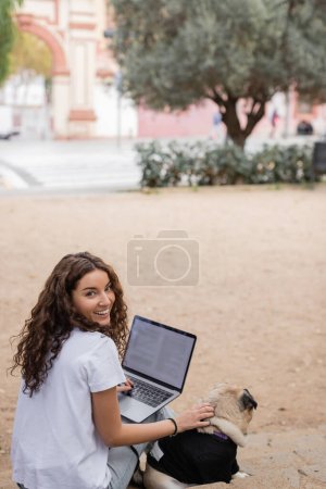 Photo for Carefree young and curly freelancer in casual clothes looking at camera while using laptop and petting pug dog on stairs in blurred park in Barcelona, Spain, white t-shirt - Royalty Free Image