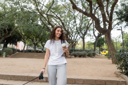 Freelancer joven y rizado sonriente con ropa casual sosteniendo café para llevar, portátil y correa mientras camina en un parque borroso durante el día en Barcelona, España 