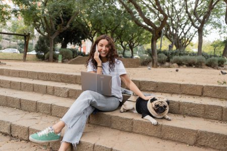 Photo for Positive and curly young freelancer talking on smartphone near laptop, looking at camera and petting pug dog while sitting on stairs in park in Barcelona, Spain - Royalty Free Image