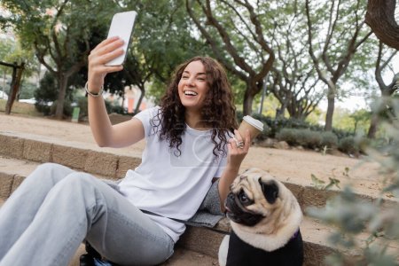 Mujer alegre joven y rizada en ropa casual que tiene videollamada en el teléfono inteligente y la celebración de un café para llevar cerca del perro pug sentado en las escaleras en el parque borroso durante el día en Barcelona, España 