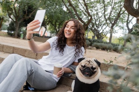 Mujer joven y feliz en ropa casual teniendo video chat en el teléfono inteligente y sosteniendo el café para ir mientras está sentado cerca de perrito en las escaleras en el parque borroso durante el día en Barcelona, España 