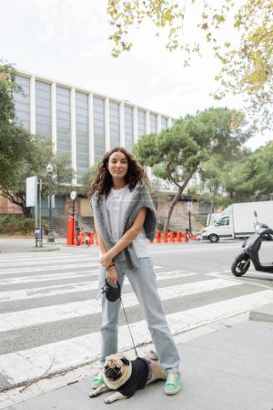 Mujer joven despreocupada con ropa casual mirando a la cámara y con correa mientras está de pie cerca de un perrito cerca del edificio en la borrosa calle urbana durante el día en Barcelona, España 