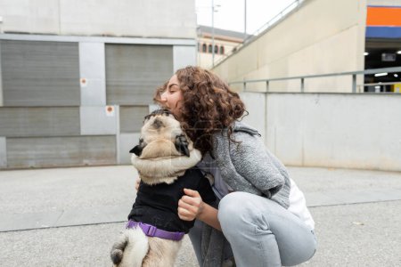 Young and curly woman in casual and soft clothes petting pug dog in sweater while spending time on blurred urban street with buildings at background in Barcelona, Spain 