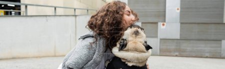 Photo for Young and brunette woman in casual clothes smiling and petting pug dog in sweater near blurred building on urban street at daytime in Barcelona, Spain, banner, having fun - Royalty Free Image