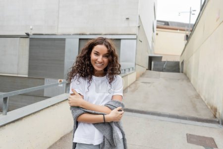 Portrait of smiling curly and pretty woman in white t-shirt and sweater looking at camera while standing near blurred industrial building at background on city street in Barcelona, Spain 