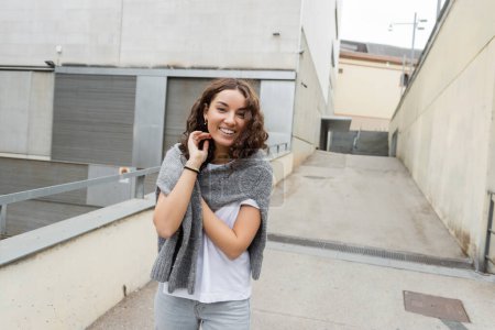 Mujer joven y rizada alegre en camiseta blanca tocando el suéter caliente y mirando a la cámara mientras está de pie cerca de un edificio industrial borroso en la calle urbana de Barcelona, España 