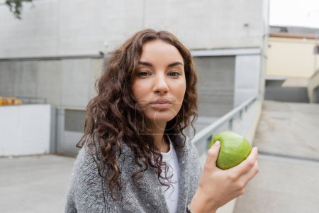 Porträt einer jungen, lockigen Frau in warmem Pullover, die einen frischen grünen Apfel in der Hand hält und bei Tag in die Kamera auf einer verschwommenen städtischen Straße in Barcelona blickt, Spanien, Industriegebäude