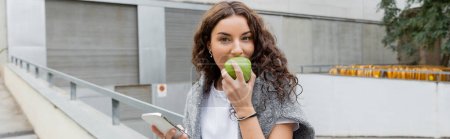 Mujer morena joven con suéter caliente en hombros comiendo manzana verde madura y sosteniendo el teléfono inteligente mientras mira la cámara en la calle urbana de Barcelona, España, pancarta, edificio industrial