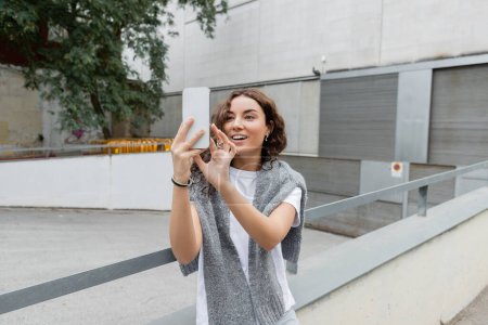 Photo for Carefree young brunette woman with warm grey sweater on shoulders having video call on smartphone while standing on urban street with industrial building at background in Barcelona, Spain - Royalty Free Image