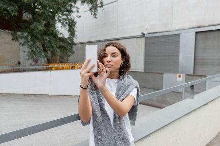 Bonita mujer morena con suéter cálido en hombros usando teléfono móvil mientras está de pie en la calle urbana con un edificio industrial borroso en el fondo en Barcelona, España 