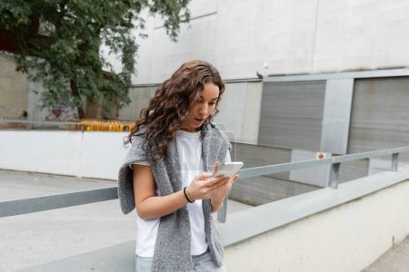 Jeune femme bouclée en pull chaud sur les épaules à l'aide d'un smartphone tout en se tenant debout sur la rue de la ville avec des bâtiments en arrière-plan pendant la journée à Barcelone, Espagne 