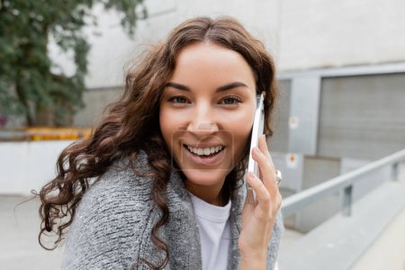Portrait of positive and young curly woman with sweater on shoulders looking at camera while talking on smartphone on blurred urban street in Barcelona, Spain 