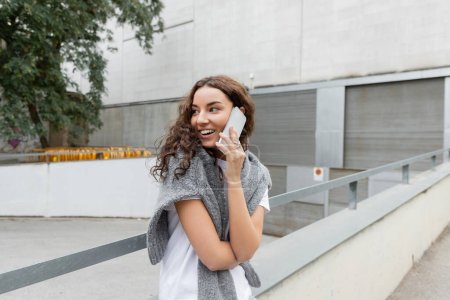 Cheerful and young curly woman in t-shirt and warm sweater talking on smartphone and looking away while standing near blurred buildings on urban street in Barcelona, Spain, green tree
