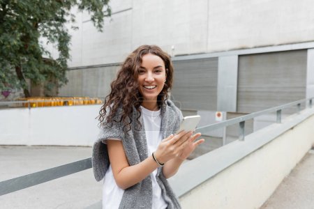 Portrait of positive and curly young woman in t-shirt and sweater looking at camera and using mobile phone while standing on city street at daytime in Barcelona, Spain, green tree