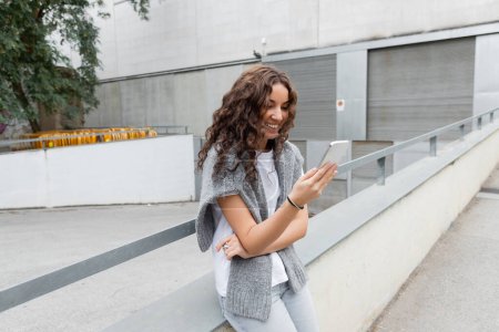 Smiling brunette woman in t-shirt with warm sweater on shoulders using smartphone while standing on blurred city street at daytime in Barcelona, Spain, green tree