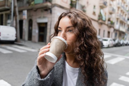 Portrait of young and curly brunette woman in casual jacket drinking coffee to go from paper cup and looking away while standing near blurred building on city street in Barcelona, Spain 