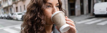 Young brunette and curly woman looking away while drinking coffee to go from paper cup and standing on blurred city street with buildings in Barcelona, Spain, banner 