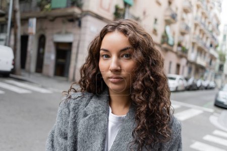 Portrait of young and curly brunette woman in casual grey jacket standing on blurred urban street with buildings at background at daytime in Barcelona, Spain 