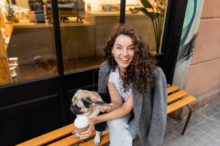 Positive junge brünette Frau in lässiger Jacke mit Smartphone und Kaffee to go, während sie neben dem Mops Hund auf einer Bank in der Nähe eines Straßencafés in Barcelona sitzt, Spanien 