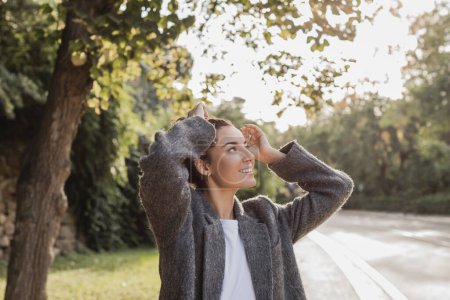 Téléchargez les photos : Jeune femme insouciante en veste grise décontractée touchant les cheveux bruns et regardant loin tout en se tenant dans un parc vert flou à Barcelone, Espagne - en image libre de droit