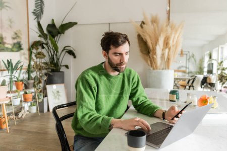 freelancer in green jumper holding smartphone and using laptop while working near coffee cup