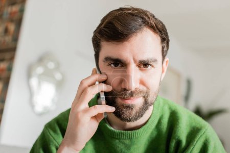 Porträt eines brünetten und bärtigen Mannes im grünen Pullover, der mit dem Smartphone spricht und in die Kamera blickt