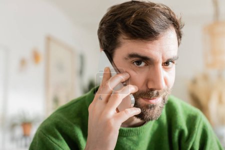 Retrato del hombre en jersey verde informal borrosa mirando a la cámara y hablando en el teléfono inteligente