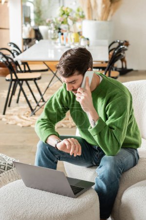 Mann in grünem Pullover und Jeans schaut auf Armbanduhr und spricht auf Smartphone, während er am Laptop arbeitet 