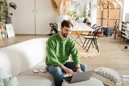Photo for Cheerful bearded freelancer in jumper and jeans working on laptop while sitting near smartphones - Royalty Free Image