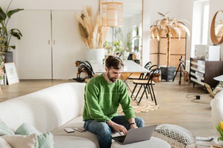 Photo for Bearded tattooed freelancer in jumper and jeans using laptop while working and sitting on couch - Royalty Free Image
