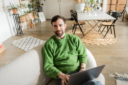 Foto de Freelancer alegre y barbudo en jersey verde usando laptop y mirando hacia otro lado, trabajo remoto - Imagen libre de derechos