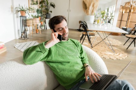 Bearded freelancer in green jumper and jeans talking on smartphone while using laptop at home 
