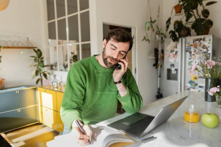 Freelance barbu en pull vert parler sur smartphone et écrire sur ordinateur portable, travail à distance