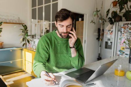Bearded freelancer having smartphone conversation and writing on notebook while working at home 