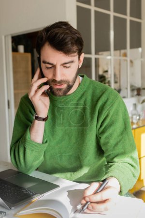 Portrait of bearded entrepreneur in green jumper talking on smartphone and writing on notebook 