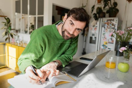 Smiling bearded freelancer in green jumper looking at camera while writing on notebook near devices