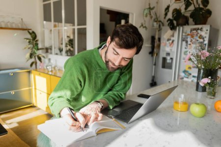 Bearded freelancer in green jumper writing on notebook while working near devices at home 